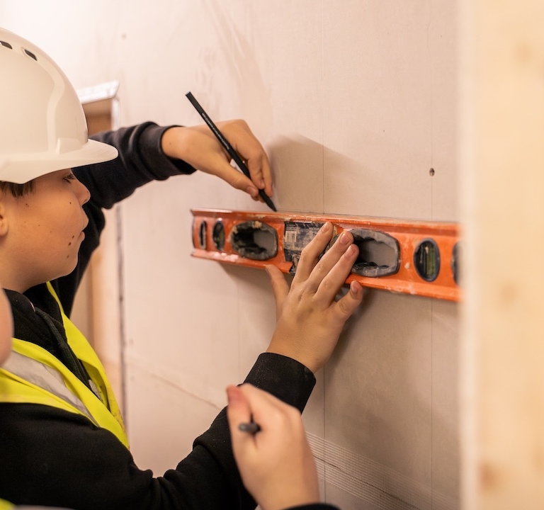 A learner using a spirit level on a wooden wall, and lining with a marker