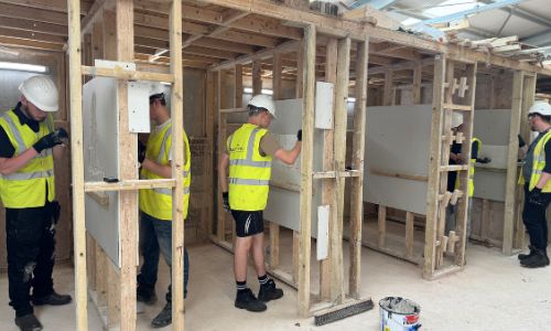 young people in high vi's jackets and hard hats, tiling a wall in a workshop setting