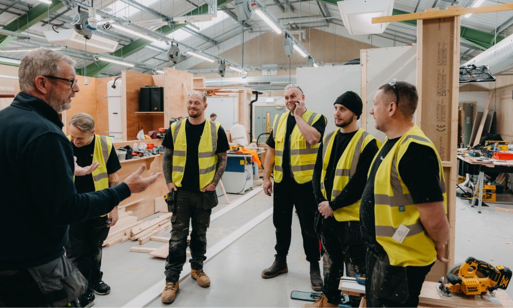 five glp training apprentices in high vi's jackets, standing listening to a teacher who is shown to be explaining. in a busy workshop setting.