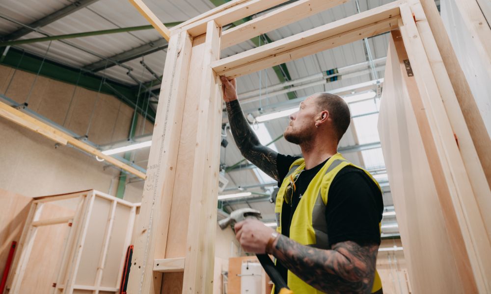 apprentice at a glp training building site, nailing a door with a hammer. The person is male, short hair, wearing a black t shirt and a high vis jacket