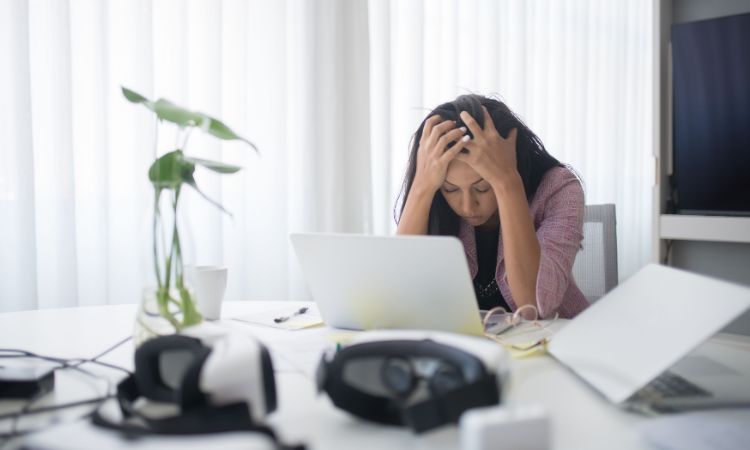 a woman on her desk, with her laptop and notes, looking stressed with her head in her hands