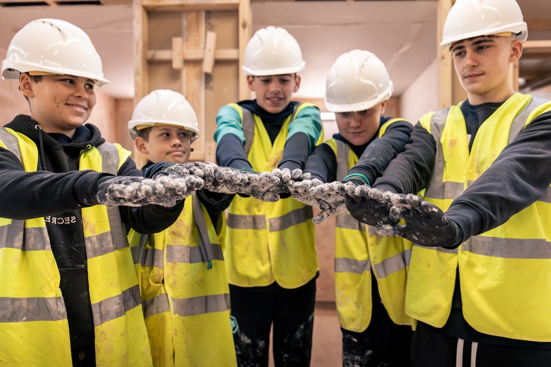Five young people in safety helmets and high-visibility vests reaching in and placing their gloved hands together.